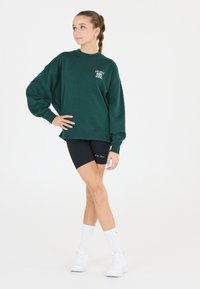 Grön oversized sweatshirt med rund halsringning och långa ärmar, matchad med svarta cykelbyxor och vita träningsskor och strumpor.