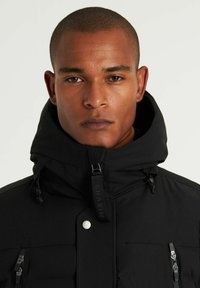 Svart vadderad jacka med hög krage, justerbara snören, dragkedjefickor och en logotags. Slät textur och minimalistiska designdetaljer.