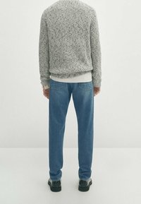 Pull en maille gris, couche légère en coton en dessous, jean en denim bleu, chaussures noires, vu de derrière sur un fond clair.