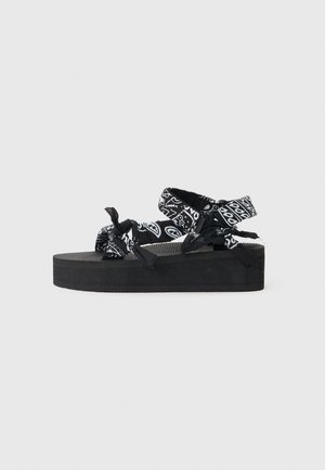 Black platform sandal with thick sole and adjustable black-and-white patterned fabric straps against a plain gray background.
