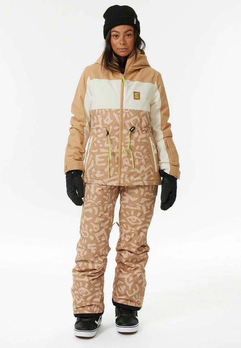 Giacca da sci isolata marrone e crema con motivi geometrici, abbinata a pantaloni marroni coordinati e guanti e stivali neri.
