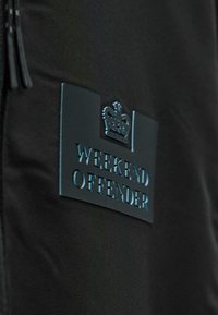 Čierna tkanina s čiernou nášivkou, na ktorej sa nachádza symbol koruny a embosovaný text "WEEKEND OFFENDER."