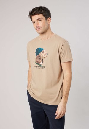 Man draagt een beige t-shirt met een afbeelding van een hond met een blauwe hoed en tekst, staat met handen in de zakken van een donkere broek tegen een effen achtergrond.
