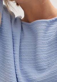 Jasnoniebieska, teksturowana tkanina z drobnymi pionowymi fałdami. Miękki, lekki materiał, który gładko opada wokół dekoltu.