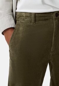 Olive green corduroy pants with a textured, ribbed surface, featuring side pockets and a button closure. Partially visible striped sleeve.