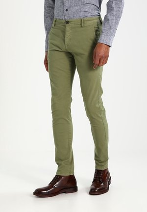 Chino verde in cotone con una vestibilità slim, dotati di chiusura a bottone, tasche anteriori e posteriori e una texture liscia. Indossati con stivali in pelle marrone.