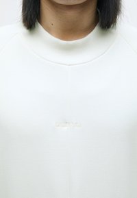 Bílá mikina s vroubkovaným kulatým výstřihem a jemně embosovaným logem "OAKLEY F.G.L." na přední straně. Hladká textura a volný střih.