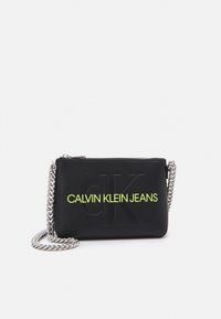 Černá kožená crossbody taška se stříbrným řetízkovým popruhem, s vyraženým logem "CK" a jasně žlutým textem "CALVIN KLEIN JEANS" na přední straně.