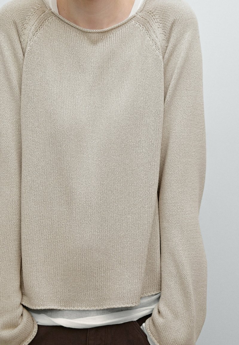 Beige gebreide trui met een ronde halslijn en raglanmouwen. Heeft een cropped ontwerp, gestructureerde stof en een gelaagd wit shirt eronder.