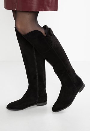 Black suede knee-high boots worn with sheer black tights and a burgundy skirt with a front zipper, shown on a white background.