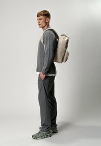 Sac à dos beige de forme rectangulaire avec plusieurs compartiments, porté par une silhouette masculine en pull gris et pantalon sombre, sur un fond neutre.