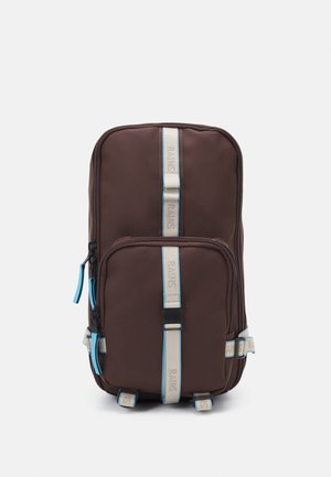 Brązowy prostokątny plecak z przednią kieszenią, beżowo-niebieskimi paskami z logo i niebieskimi suwakami.