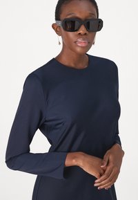 Námorně modré šaty s dlouhým rukávem z hladké tkaniny, s kulatým výstřihem. Modelka má na sobě tmavé, ostré sluneční brýle a nenápadné náušnice.