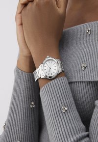 Sølv metal ur med en rund hvid urskive og sølv timermærker, vist på et håndled dækket af en ribbet grå sweater.