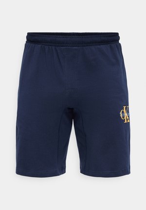 Laivastonsiniset, joustavat vyötäröshortsit, joissa on Calvin Kleinin logo keltaisena ja valkoisena oikeassa reisiosassa, esitettynä edestä valkoisella taustalla.
