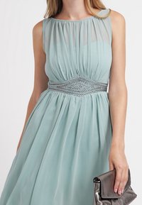 Bezramenné šaty v jemné mátově zelené barvě s nařaseným korzetem a elastickým pasem, zdobené složitými stříbrnými detaily. V doprovodu metalické kabelky.