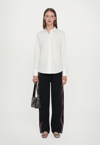 Camicia bianca a maniche lunghe con bottoni, pantaloni neri a gamba larga con strisce colorate, scarpe basse multicolori e una piccola borsa a tracolla grigia.
