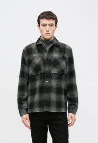 Μαύρο και γκρι πουκάμισο-τζάκετ με καρό σχέδιο, κατασκευασμένο από απαλή, υφή υφάσματος. Διαθέτει δύο τσέπες στο στήθος και μπροστινό κούμπωμα με κουμπιά, με γιακά στο λαιμό.