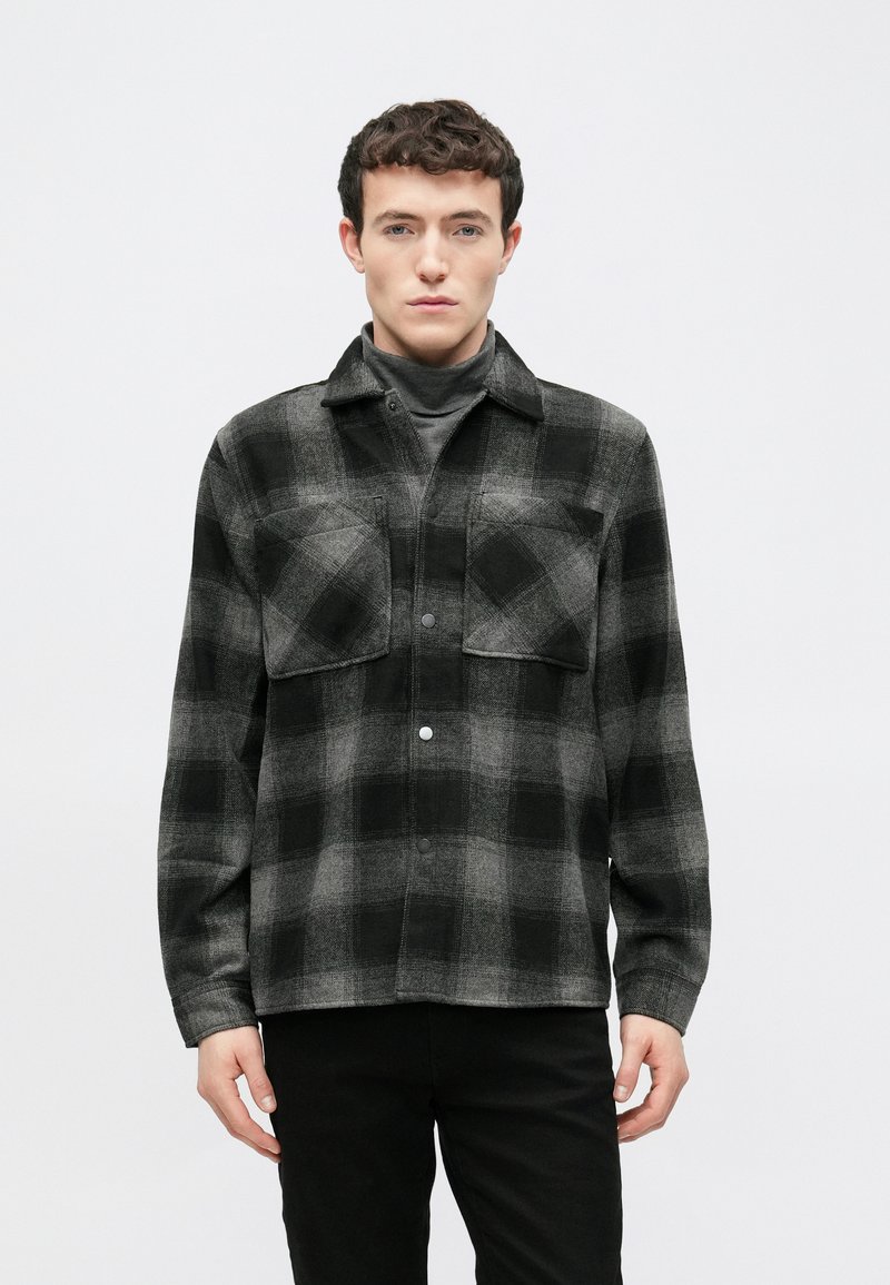 Μαύρο και γκρι πουκάμισο-τζάκετ με καρό σχέδιο, κατασκευασμένο από απαλή, υφή υφάσματος. Διαθέτει δύο τσέπες στο στήθος και μπροστινό κούμπωμα με κουμπιά, με γιακά στο λαιμό.