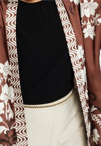 Veste à motifs floraux marron portée sur un haut noir rentré dans un pantalon crème avec une bordure décorative dorée et marron à la taille.