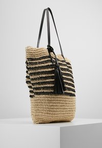 Tkaná slámová taška s černými pruhovanými akcenty, plochým dnem a detailem střapce. Má dlouhá černá ramena.