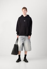 Schwarzer Hoodie mit Vordertasche und Logo, hellblaue Cargo-Shorts, schwarze Socken und schwarze Schuhe, trägt eine schwarze Tragetasche.