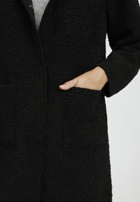 Černý texturovaný kabát s plyšovou, měkkou tkaninou, se dvěma předními kapsami a zapínáním na jeden knoflík. Design má volný střih.