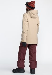 Beige jacka med huood, svarta handskar, murriga byxor med elastiska muddar och svarta stövlar. Har en enkel, funktionell design utan mönster.