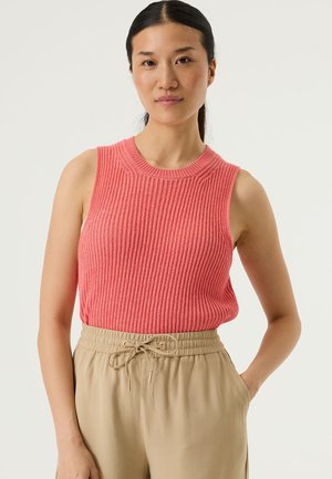 Haut sans manches en tricot couleur corail vif, avec une texture côtelée et un col rond, associé à un pantalon beige à cordon de serrage.