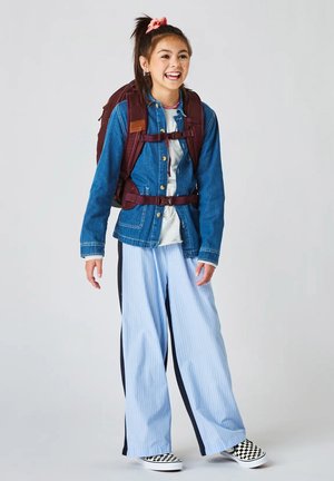 Teenager-Mädchen lächelt und trägt eine Jeansjacke, gestreifte weite Hosen, Schachbrett-Slipper und einen weinroten Rucksack mit Brust- und Taillengurten.