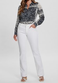 Blouse à manches longues avec un motif paisley marine et crème, associée à un pantalon blanc évasé. Le haut présente un col et une fermeture à boutons.