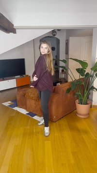 Oversized bordeauxrode trui, zwarte legging en zilveren sneakers. Getextureerde bruine bank, houten vloer en groene kamerplant op de achtergrond.