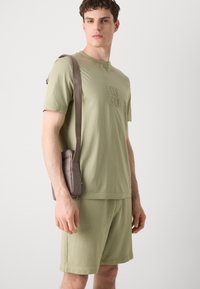 Olive green short-sleeve t-shirt with embossed "HUGO" logo, paired with matching shorts. A small brown crossbody bag with a strap is worn.