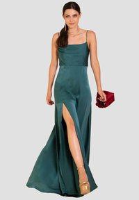 Abito in satin verde smeraldo con scollatura quadrata, spalline sottili e uno spacco alto fino alla coscia; abbinato a una pochette rossa e a tacchi alti.