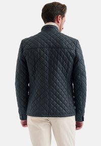 Gequiltede donkergrijze jas met een opstaande kraag, voorzien van een ruitpatroon en knoopmanchetten. Het achteraanzicht toont een getailleerde pasvorm en naaddetails.