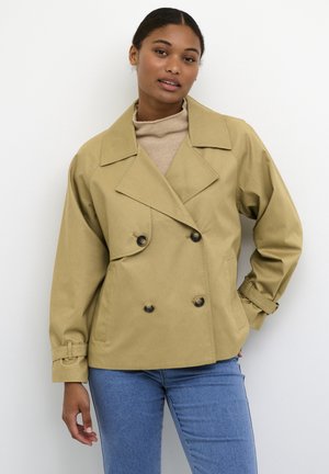 Khaki värvi kahekordne jakk suurte kraede ja nööbi aktsentidega, millel on lai lõige ja mansetiga varrukad, kantud beežil turtleneck'il.
