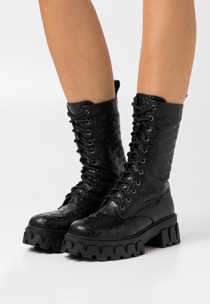 Bottes de combat noires à lacets avec motif croco texturé et semelles épaisses à crampons portées sur jambes nues sur fond blanc.