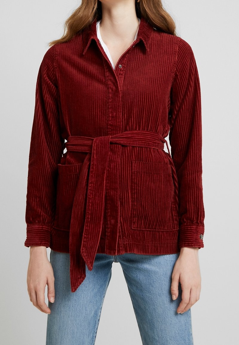 Bordeaux corduroy jack met een ceintuur in de taille, twee voorzakken en knoopdetails op de mouwen, gecombineerd met lichtblauwe jeans.