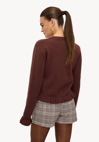 Pull bordeaux avec ourlet et poignets à volants, porté avec un short à carreaux. Texture lisse et design décontracté, idéal pour le superposition.
