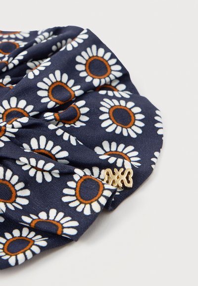 Chouchou en tissu bleu marine avec des motifs de marguerites blanches et orange et un petit charme décoratif en or attaché sur un fond blanc.