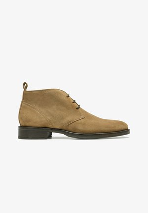 Bottines en suède tan avec un bout effilé, trois œillets et une languette à l'arrière. La semelle est sombre et légèrement texturée.