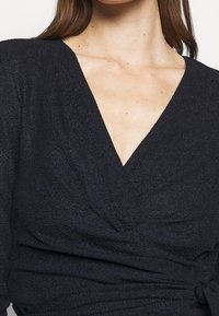 Černý hladký top s texturou, který má hluboký V-výstřih a nařasené detaily v oblasti pásu. Měkká látka s mírným leskem.