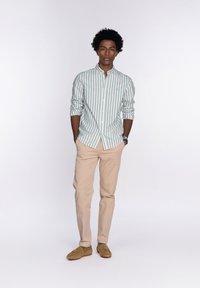 Camisa de manga larga a rayas verde y blanca, abotonada, combinada con pantalones beige y mocasines de ante. El modelo está de pie con las manos en los bolsillos.