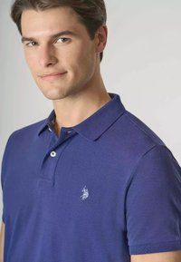 Polo shirt viola con maniche corte, colletto e un logo ricamato discreto sul lato sinistro del petto. Tessuto dalla trama liscia, colore uniforme.