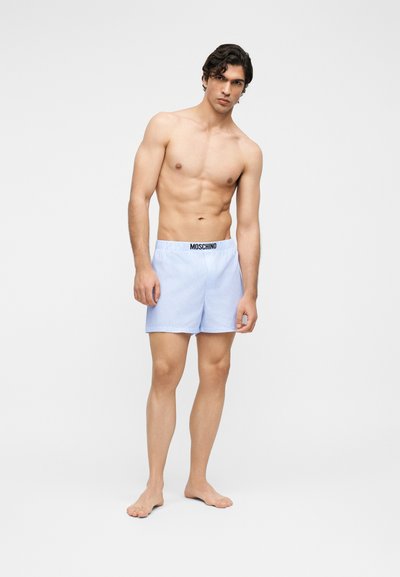 Νέος άνδρας στέκεται ξυπόλητος φορώντας ανοιχτό μπλε ριγωτά εσώρουχα Moschino μπροστά σε ένα απλό λευκό φόντο.