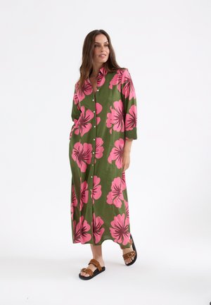 Mujer con un vestido largo verde con grandes estampados florales rosas y sandalias marrones con tachuelas, de pie frente a un fondo blanco liso.