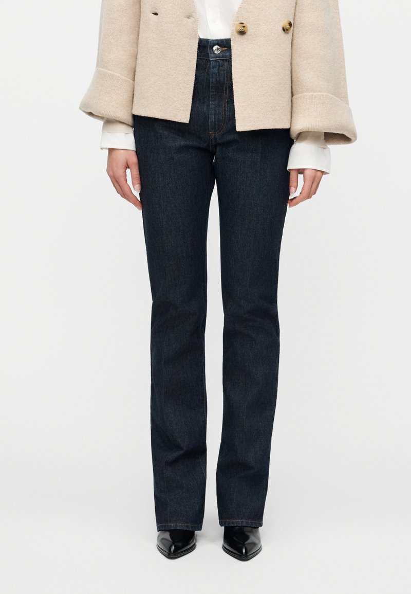 Γυναίκα που φοράει σκούρα μπλε τζι ν με ίσια μπατζάκια, μπεζ παλτό διπλής σειράς με γυρισμένα μανίκια, μάνικες λευκού πουκαμίσου και μαύρα μυτερά παπούτσια.