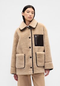 Giacca in pelliccia sintetica beige con rifiniture nere, grandi tasche frontali e tasca sul petto con zip. Presenta un colletto arrotondato e chiusura con bottoni.