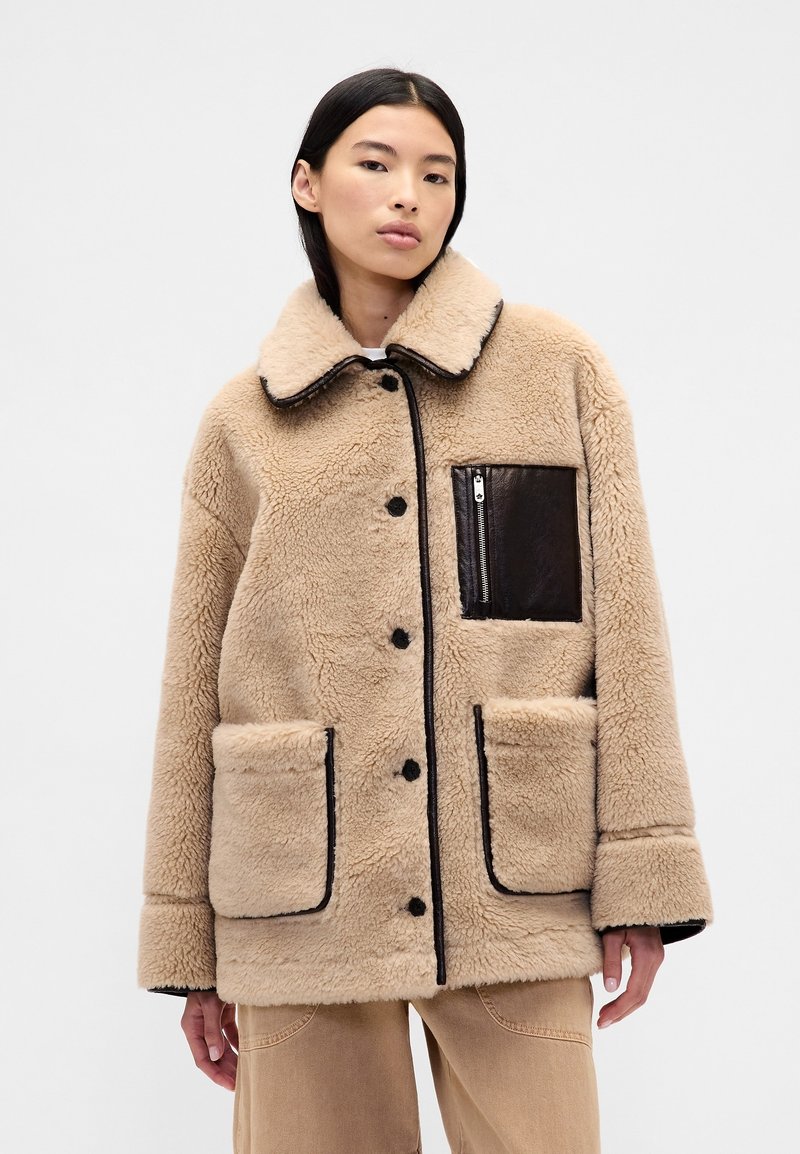 Giacca in pelliccia sintetica beige con rifiniture nere, grandi tasche frontali e tasca sul petto con zip. Presenta un colletto arrotondato e chiusura con bottoni.