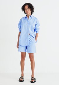 Chemise en coton bleu clair avec un devant à boutons, assortie à un short. Modèle portant des sandales noires, debout devant un fond blanc.